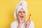 Sad man wrapped towel on head doing morning cosmetology procedures, posing with patches under eyes isolated over yellow background
