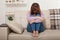 Sad middle aged woman sitting on sofa with clamped knees and crying. Home background