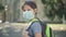 Sad schoolboy with backpack in face mask turning to camera. Portrait of upset Caucasian boy standing on sunny schoolyard