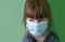 A sad small girl in a medical mask on a green background. Disposable medical mask on the child`s face. A child in a protective med