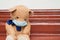 Sad teddy bear toy wearing disposable medical protective face mask, sitting on vintage brown bench. Stay at home and