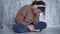 Sad teenage girl sits alone near a gray wall, bending over her smartphone. Schoolgirl wears headphones and listens to