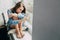 A sad teenager girl in a blue shirt sits near the window on the pillow and looks at the smartphone and chat with friends, hugging