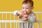 Sad toddler baby boy in the crib gnawing a wooden toy, yellow studio background. A tired child in pajamas