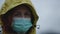 Sad woman in green antivirus face mask and yellow hood looks at you and moves eyes away during coronavirus epidemic