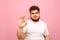 Sad young man in a white T-shirt and overweight stands on a pink background, looks at the camera and shows an OK gesture. Upset