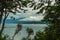 Sailboat navigating a calm lake with low clouds and mountains in the background. View between the trees. Villa La Angostura,