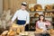 The saleswoman starts cutting the bread and the baker arranges the rolls and other goods in the store. Family bakery and
