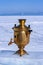 A samovar stands on a piece of ice on a blurry background of a lake in the snow and mountains in the distance.