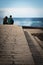 San Sebastian, Spain - March 16, 2018: couple sitting on stone wall admiring scenic huge splash of sea water waves