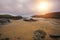 A sandy bay with scattered rocks opening to the sea, with a bright sun filtering through a pink, cloudy sky