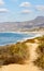 Sandy pathway lined with weeds and wildflowers with hilltop view of ocean shoreline