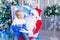 Santa Claus holds a baby in his arms near a Christmas tree in a blue Christmas interior, a happy child talks to Santa Claus