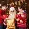 Santa Claus opens a bag and gives gifts to children, sitting indoor near decorated xmas tree with lights - Merry Christmas and