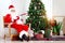 Santa Claus in red costume playing Ukulele and singing Christmas songs front of decorative Christmas tree in living room. Happy