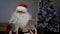 Santa Claus sits on couch with a tablet and prepares gifts for children and adults. Santa Claus is working, preparing a