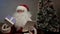 Santa Claus sits on couch with a tablet and prepares gifts for children and adults. Santa Claus is working, preparing a