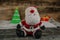 Santa doll on a wooden floor with a Christmas tree in the background and two gift boxes in front of a wooden wall