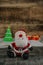 Santa doll on a wooden floor with a Christmas tree in the background and two gift boxes in front of a wooden wall