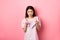 Sassy asian woman pointing at camera, smiling and inviting you, beckon or praise person, standing on pink background