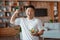 Satisfied asian mature man holding bowl with fresh salad and showing biceps, resting after domestic workout