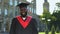 Satisfied black male in graduation gown smiling camera on university background