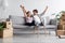 Satisfied caucasian millennial couple sit on sofa, rest, raise their hands up and celebrate purchase of new apartment