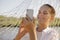 Satisfied Caucasian woman wearing white shirt sitting in hammock on the bank of the river, holding smart phone in hands, looking