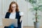 Satisfied European female with ginger hair, types information on laptop computer, has glad facial expression, drinks coffee, wears