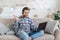 Satisfied millennial caucasian man with stubble sits on sofa waving hand and looking at laptop