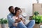 Satisfied young black husband and wife hugging, sit on floor with cardboard boxes, things and plants in new flat