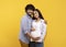 Satisfied young indian man hugging pregnant wife and touching belly, smiling together at camera over yellow background