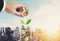 Saving money concepts, businessman hand putting coin in glass jar container, with plant bud glowing, on Bangkok city in sunrise