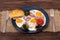 Savoury breakfast with fried eggs, bacon, tomato, broccoli and toasts in a dark plate that is on burlap, with fork and knife