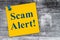Scam Alert message on a yellow sticky note on weathered wood