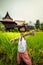 A scarecrow wearing Thai clothes in a rice paddy with Thai house in the background.