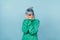 Scared hipster girl in a green sweatshirt and with blue hair stands on a blue background and covers her face in fear, looking at