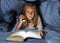 Scared little girl reading scary book under bed cover holding a flashlight late at night