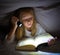 Scared little girl reading scary book under bed cover holding a flashlight late at night