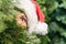 Scared, open-eyed look of a beautiful girl in a santa claus hat hiding behind a christmas tree. Concept.