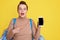 Scared pregnant woman showing phone with blank screen, pointing at smartphone with index finger, keeps mouth widely opened, feels
