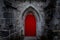 Scary pointy red wooden door in an old and wet stone wall building with cross, skull and bones at both sides. Concept mystery,