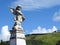 Scene in a graveyard: an old stone statue of a little angel. In the background, blue sky, clouds and vegetation.