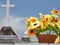 Scene in a graveyard: a vase of big fake yellow flowers. In the background, a blurred cross.