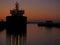 Scene at a port at sunrise. Small cargo ship silhouette with lights on. Orange and purple tones