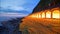 Scenery of a coastal highway by rocky beach before sunrise with lights from the rock shed tunnel illuminating the coast