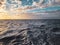 Scenic shot of turbulent waves of the blue sea under the clouds and sunset on the horizon