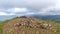 A scenic view of a rocky mountain summit with a stony cairn and mountain range and valley in the background under a stormy grey