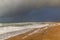 A scenic view of a sandy beach with crashing waves, rocky groyne under a dark black stormy sky