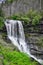 A scenic waterfall roadside going over the cliff down to the boulders and rocks below where you can no longer drive under but can